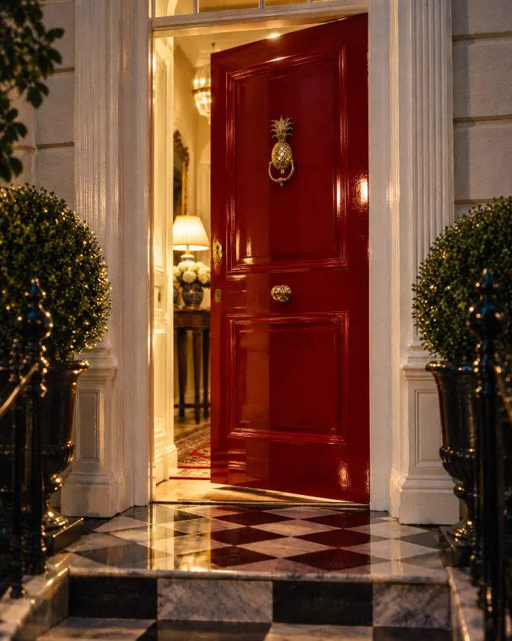 A red front door with a brass pineapple knocker, open onto a lit hallway
